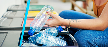 Pessoa depositando garrafas plásticas em uma lixeira de reciclagem.