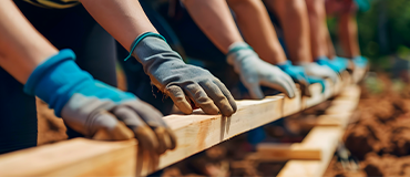 Grupo de pessoas usando luvas e levantando uma viga de madeira em um trabalho conjunto ao ar livre.