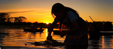 Pessoa ajoelhada na água durante o pôr do sol, mexendo em um objeto próximo à superfície.