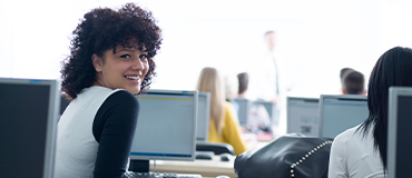 Pessoa sentada em sala de aula com computadores, olhando para a frente.